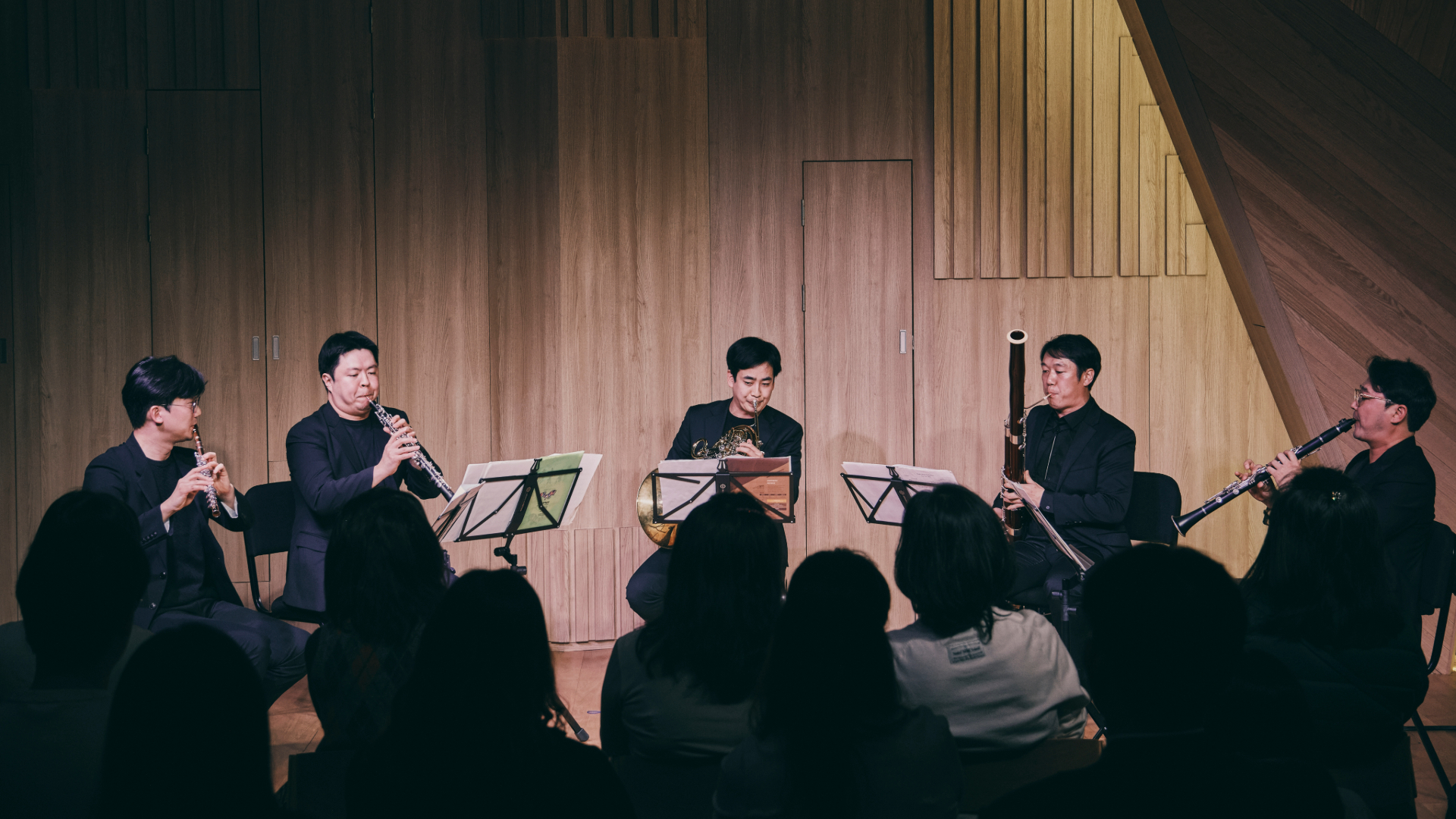플루트, 오보에, 호른, 바순, 클라리넷 연주자 다섯 명이 목관 오중주 공연을 하고 있으며, 객석의 관객들이 이를 감상하고 있다.