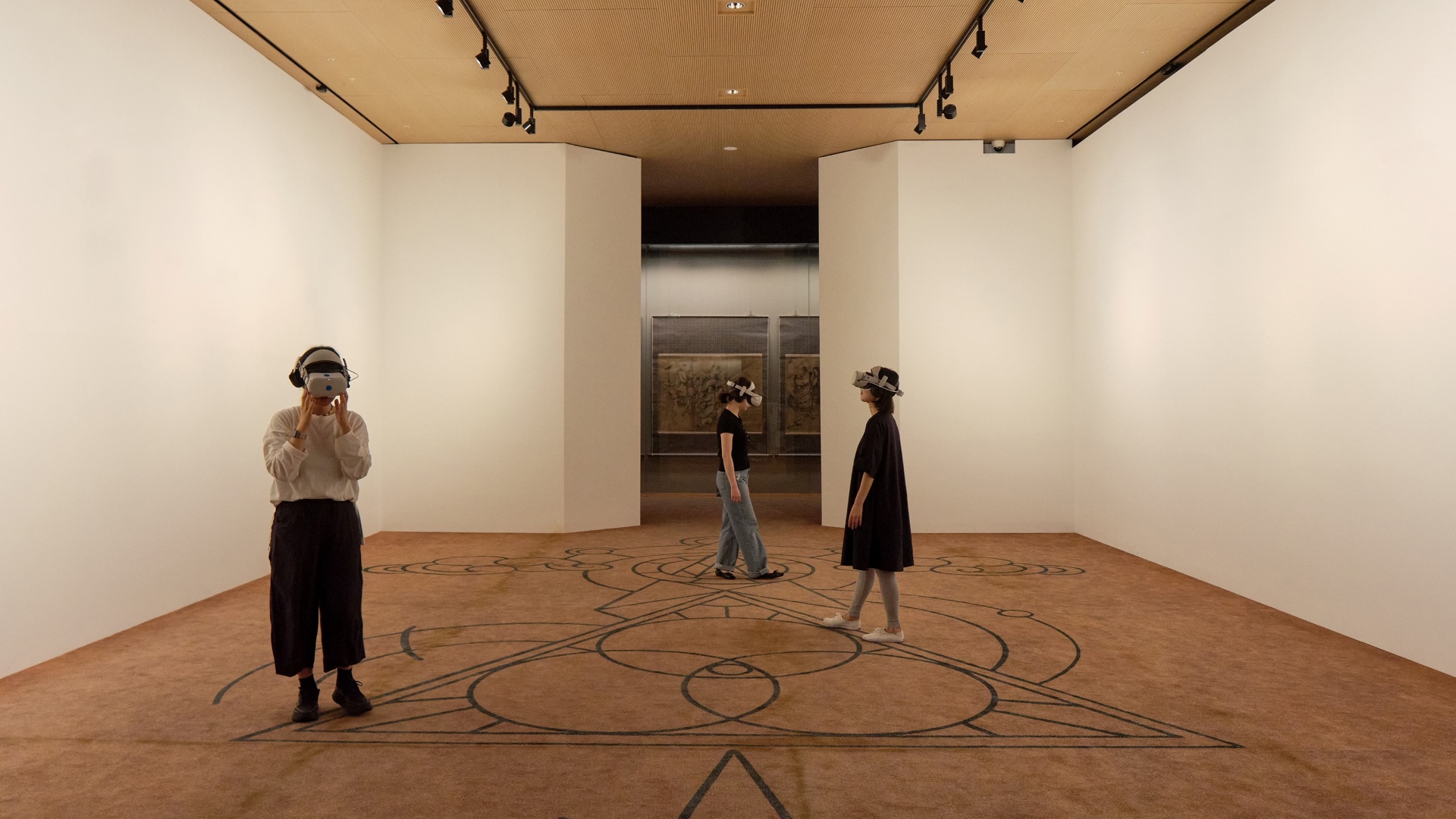 기하학적 무늬가 그려진 전시장 바닥 위에서 VR 헤드셋을 착용한 관람객 세 명이 체험 중인 모습
