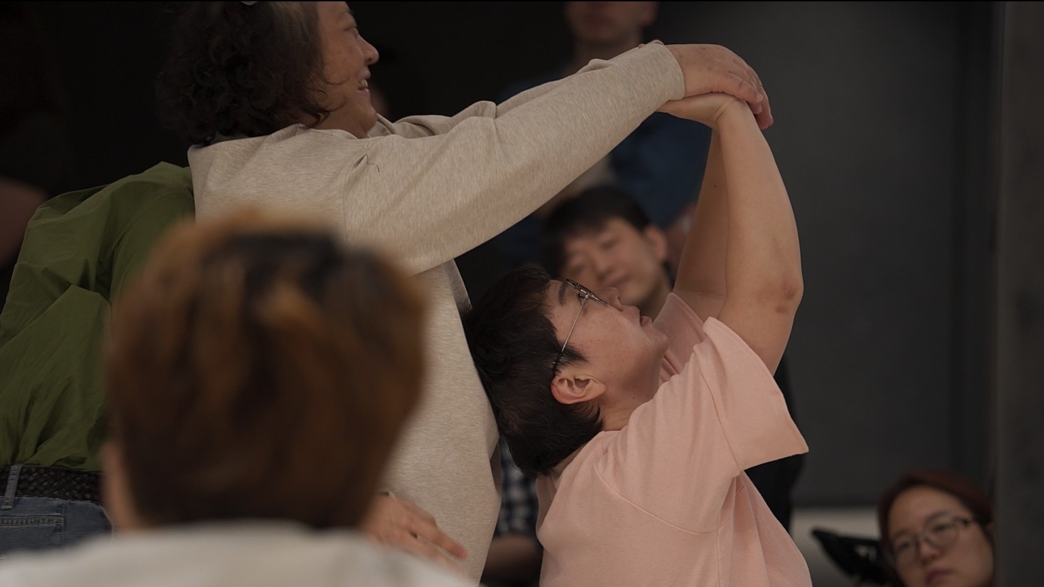 실내 워크숍 또는 퍼포먼스 현장에서 한 참가자가 다른 사람의 팔을 들어 올리며 신체 움직임을 함께 만들어가는 장면. 분홍색 티셔츠를 입고 안경을 쓴 인물이 상체를 뒤로 젖힌 채 팔이 위로 뻗어 있고, 옆의 사람이 그 팔을 부드럽게 잡고 동작을 유도하고 있다. 주변에는 여러 명의 사람들이 앉아 이 장면을 지켜보고 있으며, 일부는 흐릿하게 보이고 일부는 집중한 표정으로 관찰하고 있다.