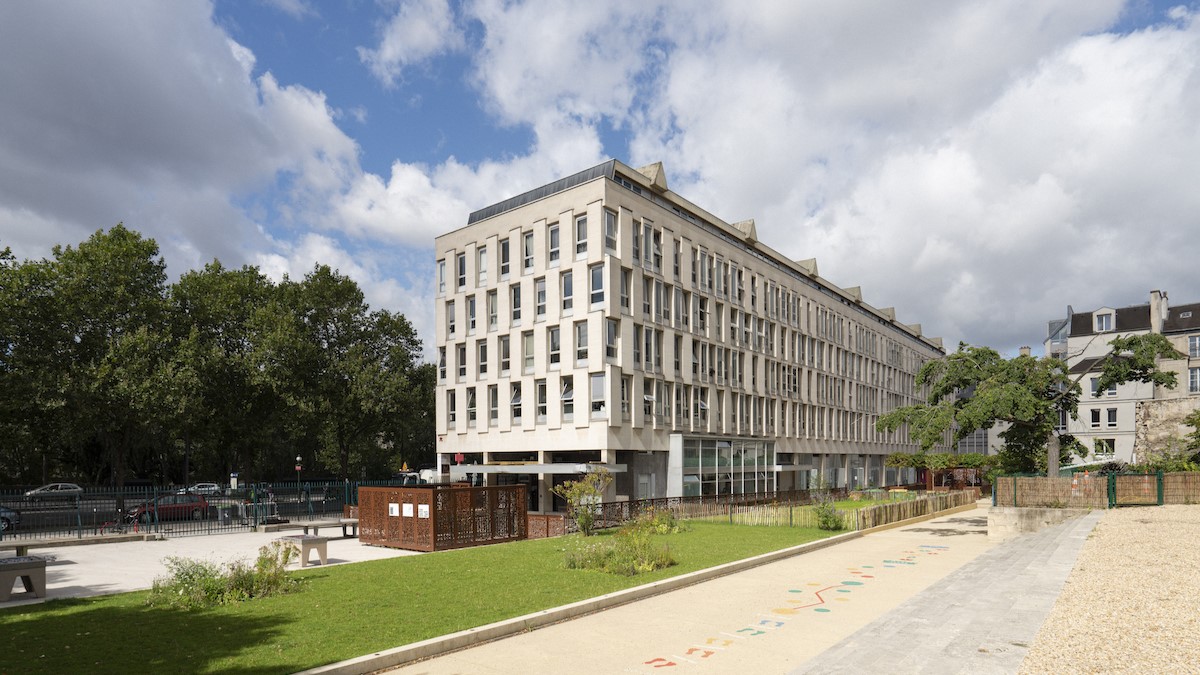 밝은 베이지색 외벽과 반복된 창문 구조를 지닌 현대식 건물. 전면에는 푸른 잔디밭과 벤치, 놀이 요소가 포함된 공공 공간이 펼쳐져 있음
