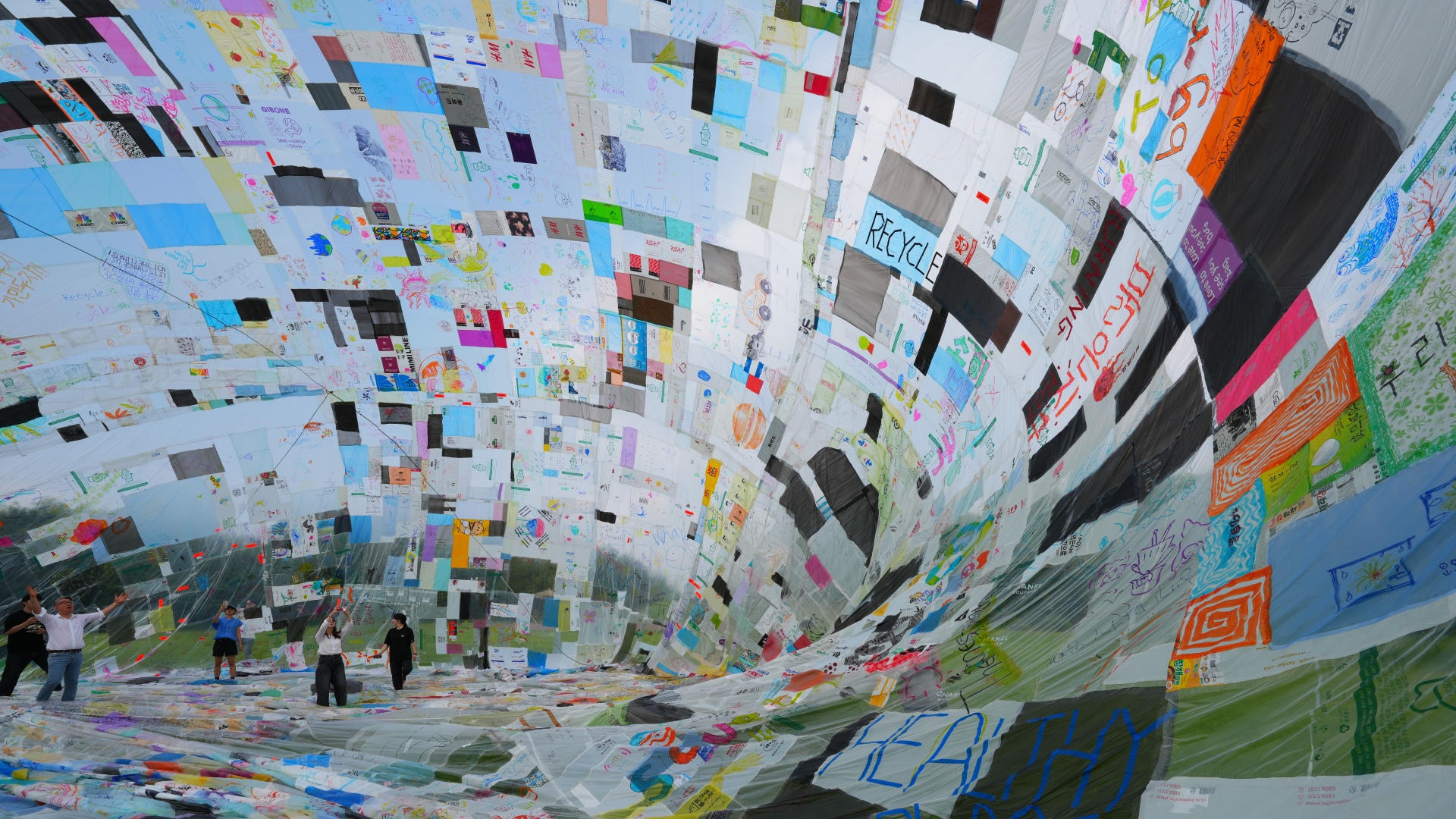 다양한 색상과 그림, 문구가 담긴 재활용 비닐 조각들을 이어 붙여 만든 대형 비닐 구조물 내부에서 사람들이 함께 들어 올리고 있는 모습