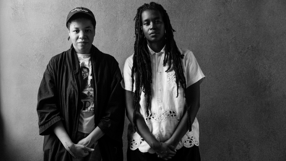 회색 배경 앞에 서 있는 두 사람의 흑백 초상사진. 왼쪽 인물은 모자와 검은 겉옷을 걸치고 있고, 오른쪽 인물은 긴 드레드 헤어스타일에 밝은 셔츠를 입고 있다